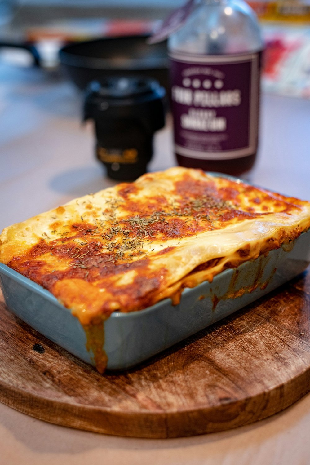 Baked lasagna in a blue dish with melted cheese and herbs on top, placed on a wooden cutting board, with a coffee cup and beverage bottle in the background.