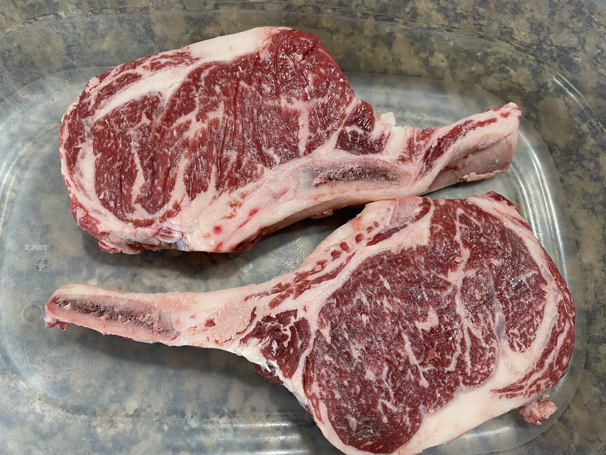 Two uncooked, marbled ribeye steaks displayed in a clear glass dish, showcasing rich red meat and white fat, ideal for grilling or roasting.