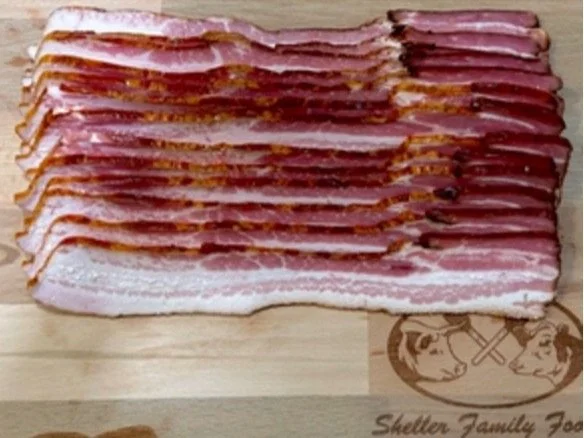 Sliced bacon arranged neatly on a wooden cutting board, showcasing its crispy texture and rich color. The board features a logo of Sheller Family Foods.