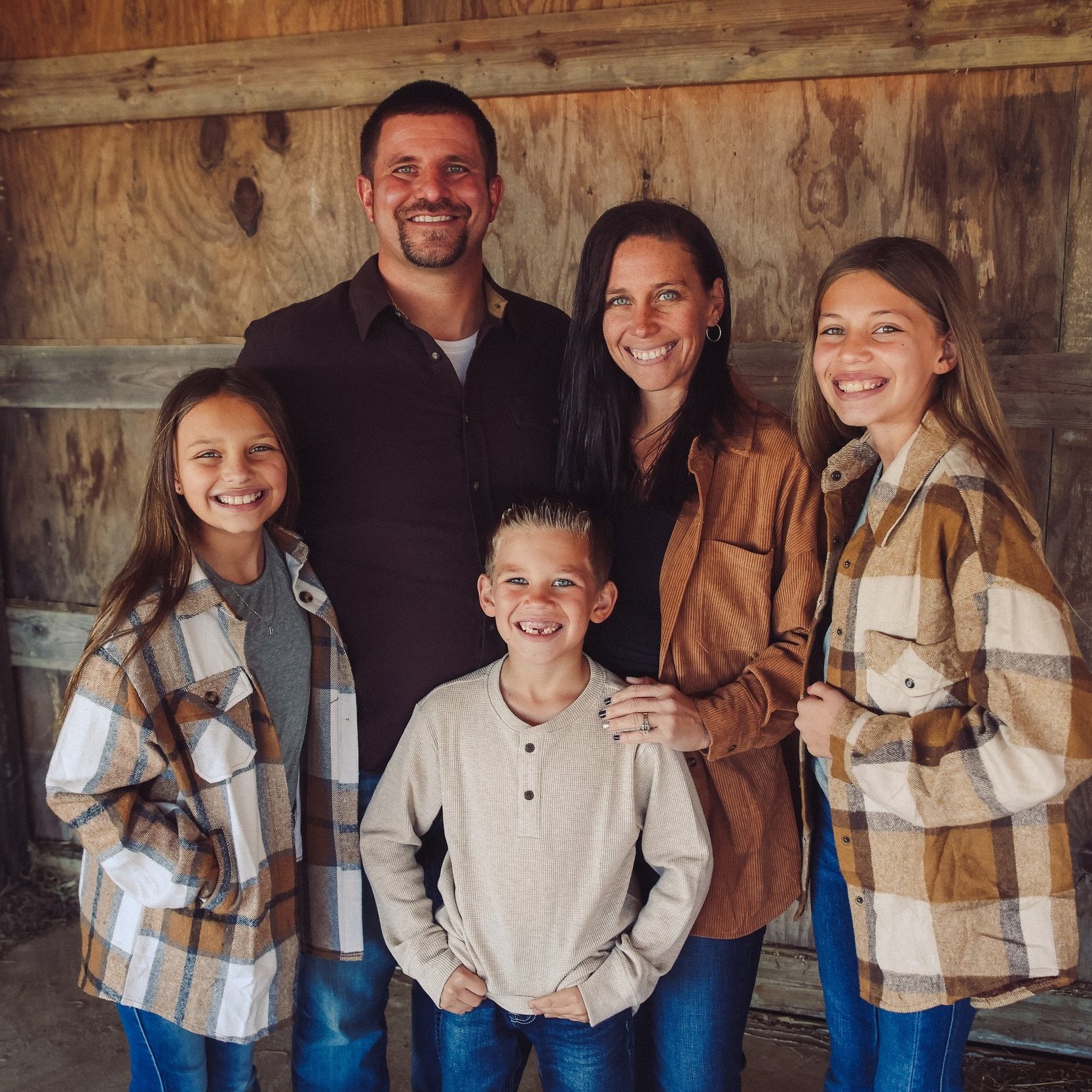 A smiling family of five poses together in a rustic setting, featuring two daughters, a son, and their parents. The children wear stylish plaid shirts, while the parents are dressed casually. The warm atmosphere highlights their joyful expressions and close-knit bond.