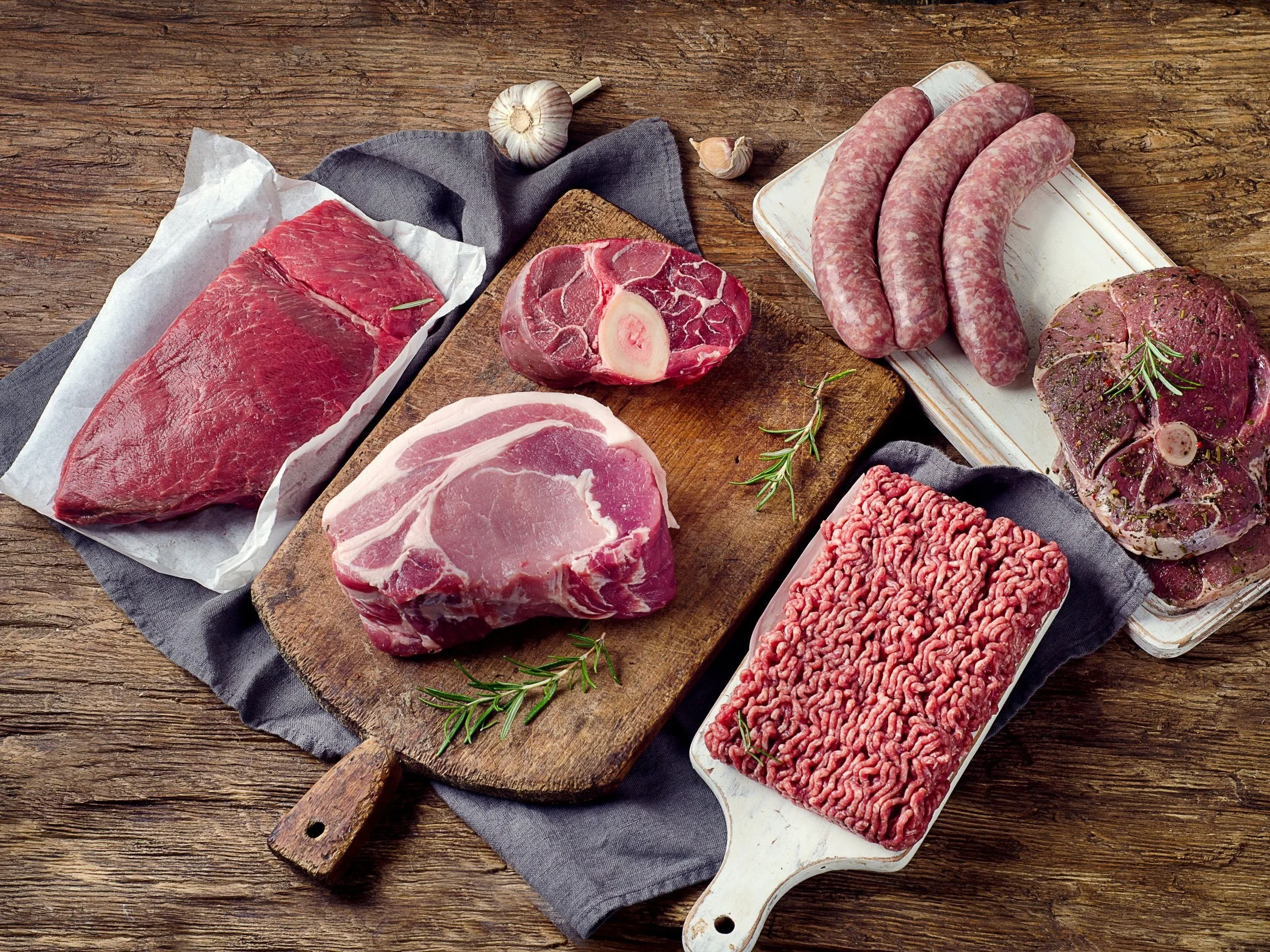 An assortment of fresh meats displayed on a wooden surface, including a beef cut, pork chop, ground beef, sausages, and a beef shank, garnished with rosemary and garlic, ideal for culinary preparation and meal planning.