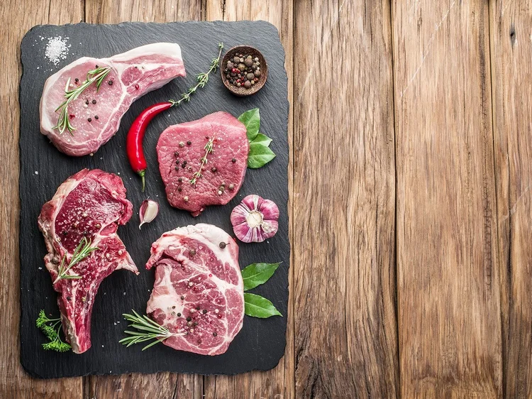 Assorted cuts of fresh meat including pork chops and beef steaks, garnished with herbs, garlic, and spices on a slate board, set against a rustic wooden background.