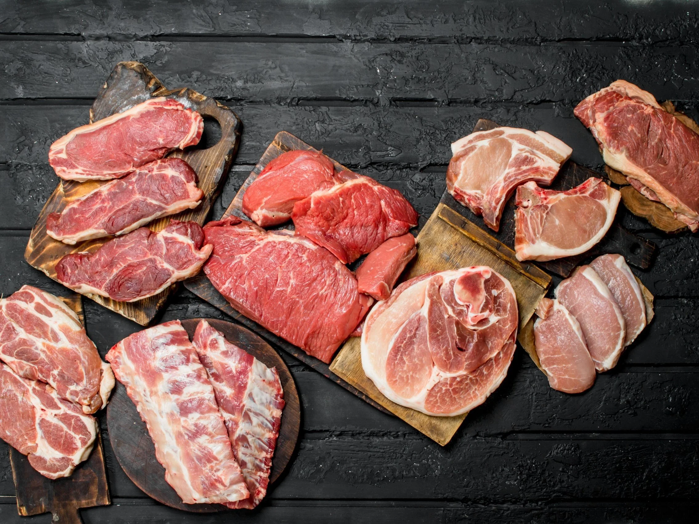Assorted cuts of raw meat displayed on wooden boards, including beef steaks, pork chops, and ribs, set against a dark wooden background. Ideal for meat lovers and culinary enthusiasts looking for fresh meat options.